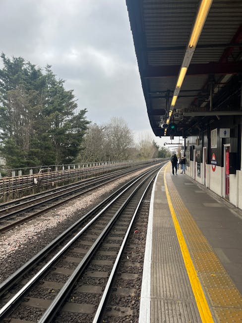 The image shows a train station platform adjacent to railway tracks extending into the distance, with a few passengers walking along the platform. The platform features a yellow tactile strip for safe
